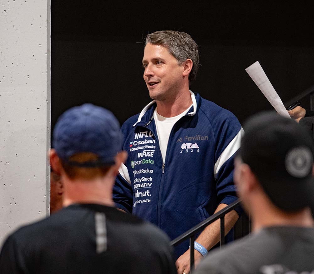 Aaron wearing a Pavilion themed jacket full of sponsor logos while speaking to a crowd of people.